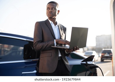 Black Handsome Businessman At Charging Station, Standing Near Electric Car With Laptop Pc And Looking Away, While Refueling His Auto. EV Station And People Concept