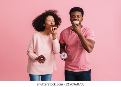 Black Happy Man And Woman Having Fun And Eating Donuts Isolated Over Pink Background