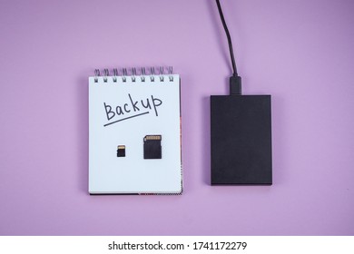 black hard drive, notepad with backup text is lying on a lilac background. Two sd cards are on the Backup concept sheet. the view from the top.