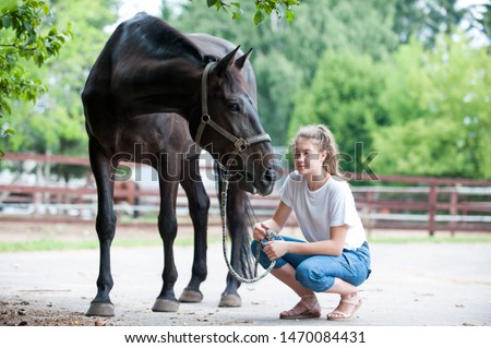 Black horse gazing away close to her owner - young teenage girl at ranch after training. Vibrant multicolored summertime outdoors horizontal image.