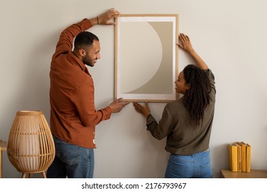 Black Husband And Wife Hanging Picture In Frame On Wall At Home. Married Couple Decorating Room With Poster Together. Interior Design, Art And Decoration Concept