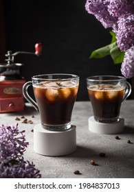 Black Iced Coffee in glass mugs with a round white plaster base. Iced coffee on a gray table. In the background are lilac flowers, a coffee grinder and coffee beans. Dark background.