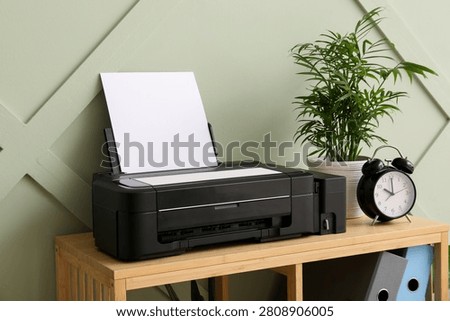 Black inkjet printer with blank paper on a wooden shelf next to a potted plant and alarm clock, the background features a sage green wall with a subtle geometric pattern
