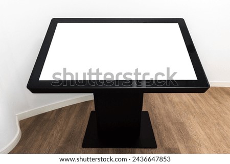 Black interactive touch table with empty blank screen stands in white interior on wooden floor, high-tech mockup