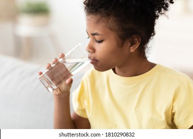 Black Kid Girl Drinking Water From Glass Sitting On Sofa At Home. Healthy Liquid Concept