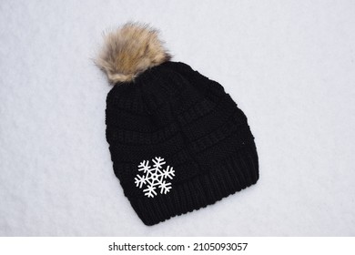 Black Knit Hat With Ball And Large White Snowflake On Fresh Snow. 