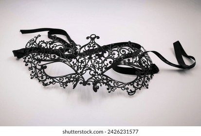 A black lace mask rests on a white surface, reminiscent of a mysterious event or a fictional character waiting to make an appearance with its intricate pattern resembling a wing or insect wing - Powered by Shutterstock