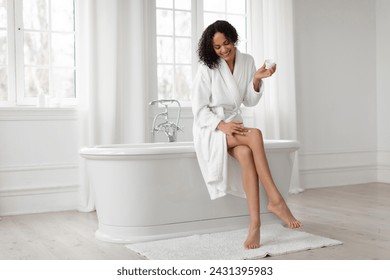Black lady in bathrobe moisturizing smooth legs with moisturizer cream after morning shower, sitting on bathtub in light bathroom. Beauty, skincare and pampering concept
