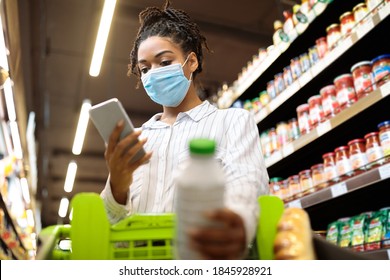 Black Lady In Mask Using Phone Shopping Buying Food Groceries Standing Wih Trolley Cart In Supermarket. Woman With Smartphone Choosing Goods In Grocery Shop Indoor. Selective Focus