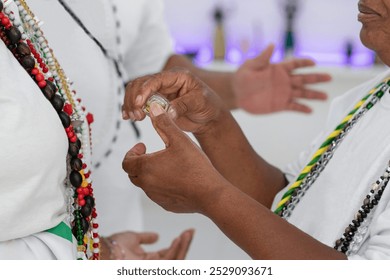 Black lady with a string of colored beads blessing a black woman with consecrated oil. In the background, a black man with a string of beads helps by sending energy and good vibrations