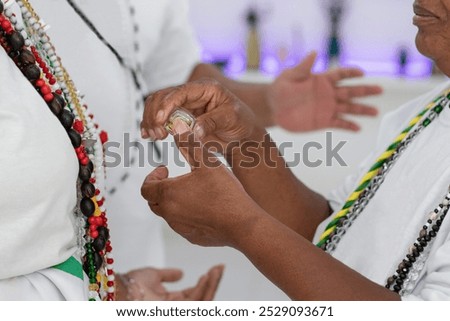 Black lady with a string of colored beads blessing a black woman with consecrated oil. In the background, a black man with a string of beads helps by sending energy and good vibrations