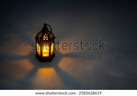 A black lantern with intricate patterns illuminates a candle against a dark background, casting a warm glow and a shadow. Copy space for text. Ramadan Kareem concept.
