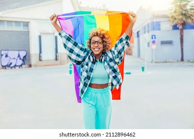 Black Latin American Woman With Eyeglasses, Headsets And Afro Hair Holds Up The Rainbow Flag Of Gay Pride That Moves With The Wind On A Sunny Day. She Fights For Sexual Freedom.