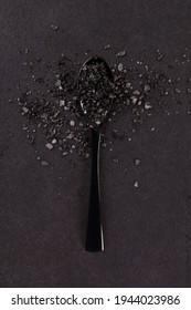 Black lava salt in black spoon on black background, top view. Culinary gourmet cuisine. 