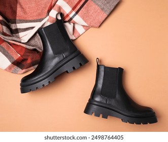 Black leather classic women's boots and checkered autumn wool scarf on beige background top view flat lay. Minimalistic layout, autumn footwear advertisement. Fashionable stylish boots