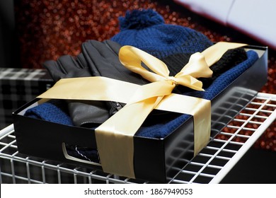 Black Leather Gloves And A Blue Knitted Hat With A Scarf Are Placed In A Dark Gift Box With A Gold Bow Side View