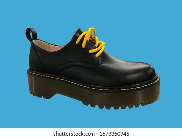 Black Leather Platform Shoes Isolated On Blue Background. Classic Shoes On A High Black Tractor Platform. Fashionable Strict Low-heeled Shoes. Women Loafers With Lacing. Composition Of Clothes