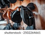 black leather saddle hanging on wooden wall at stable or barn, professional equestrian horse riding equipment