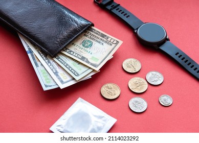 black leather wallet with dollar bills in different denominations, coins, black watch and condom on a red background