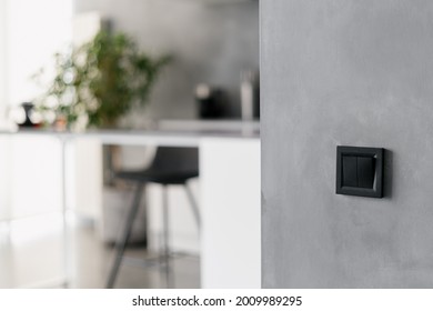 Black light switch on grey wall in modern themed kitchen environment, black bar stool, various home appliances, different white furniture and potted plant in blurred background