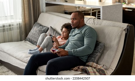 Black Little Boy And Father Playing Video Game With Joysticks On Sofa At Modern Home. Smiling Child And Focused Man Spending Time Together. Fatherhood And Parenting. Domestic Leisure And Entertainment