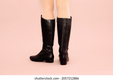 Black long autumn winter leather boots with line of rhinestones and low heels and caucasian woman legs on light powdery pink background. Studio shot photo, footwear concept. Back front view