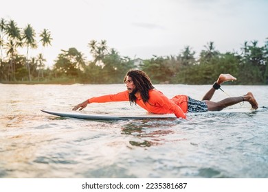 Black long-haired man paddling on long surfboard to the surfing spot in Indian ocean. Palm grove litted sunset rays in the background. Extreme water sports and traveling to exotic countries concept. - Shutterstock ID 2235381687