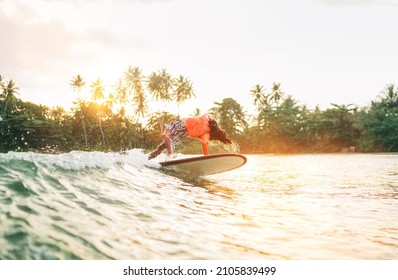 Black Long-haired Teen Boy Riding And Jumping From A Long Surfboard. He Caught A  Wave In An Indian Ocean Bay With Magic Sunset Background. Extreme Water Sports And Exotic Countries Concept.