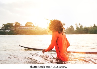 Black Long-haired Teen Boy With A Surfboard Ready For Surfing. He Walking Into Indian Ocean Waves With Magic Sunset Background. Extreme Water Sports And Exotic Countries Concept.