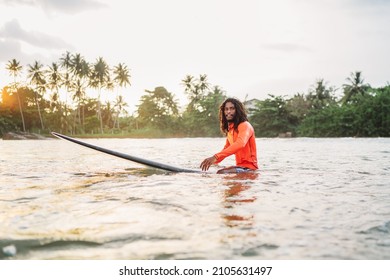 Black Long-haired Teen Man Floating On Long Surfboard, Waiting For A Wave Ready For Surfing With Palm Grove Litted Sunset Rays. Extreme Water Sports And Traveling To Exotic Countries Concept.