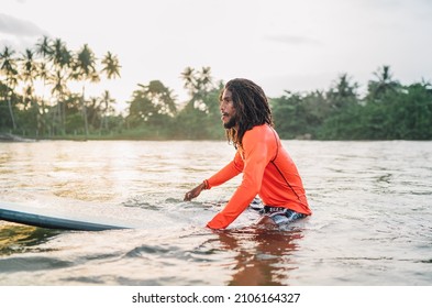 Black Long-haired Teen Man Floating On Long Surfboard, Waiting For A Wave Ready For Surfing With Palm Grove Litted Sunset Rays. Extreme Water Sports And Traveling To Exotic Countries Concept.