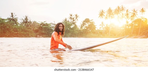 Black Long-haired Teen Man Floating On Long Surfboard, Waiting For A Wave Ready For Surfing With Palm Grove Litted Sunset Rays. Extreme Water Sports And Traveling To Exotic Countries Concept.