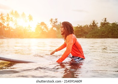 Black Long-haired Teen Man Floating On Long Surfboard, Waiting For A Wave Ready For Surfing With Palm Grove Litted Sunset Rays. Extreme Water Sports And Traveling To Exotic Countries Concept.