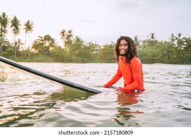 Black Long-haired Teen Man Floating On Long Surfboard, Waiting For A Wave Ready For Surfing With Palm Grove Litted Sunset Rays. Extreme Water Sports Or Traveling To Exotic Countries Concept. Sri Lanka