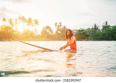 Black Long-haired Teen Man Floating On Long Surfboard, Waiting For A Wave Ready For Surfing With Palm Grove Litted Sunset Rays. Extreme Water Sports And Traveling To Exotic Countries Concept.