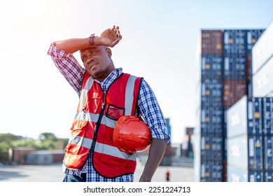 Black male African American engineering in uniform holding hard hat standing containers yard, worker tired industry frustration. Area logistics import export and shipping. - Powered by Shutterstock