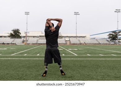 A Black male athlete with prosthetic legs stretches on football field. The athlete man prepares for sport exerice training, with determination. The athlete's prosthetics visible on sport grass field. - Powered by Shutterstock