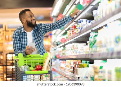 Black Male Buyer Shopping Groceries In Supermarket Taking Dairy Product From Shelf Standing With Shop Cart Indoors. Guy Buys Grocery Choosing Food In Super Market. Empty Space - Shutterstock ID 1906390078