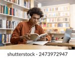 Black male college, high school student wearing headphones, sitting at desk with laptop for school project at a library interior