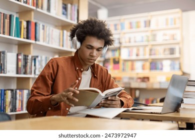 Black male college, high school student wearing headphones, sitting at desk with laptop for school project at a library interior - Powered by Shutterstock