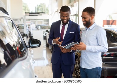 Black Male Customer In Car Dealership Center Looking At Brochure With Salesman, Happy African American Man And Salesperson Checking Vehicle Models And Characteristics In Catalog While Choosing Auto