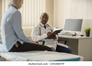 Black Male Doctor Sitting At Desk, Asking Questions To Unrecognizable Female Patient Sitting On Couch And Making Notes In Her Medical History