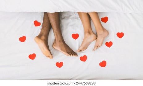 Black male and female feet lying on bed under blanket, decorated with small red paper hearts, top view - Powered by Shutterstock