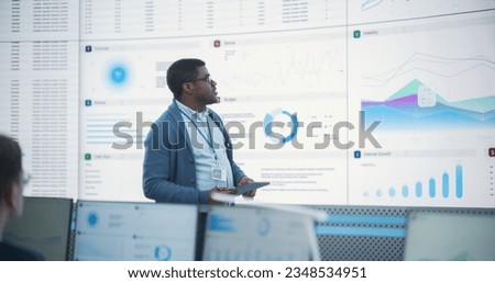 Black Male General Manager Giving Presentation To Consulting Company Employees On Big Digital Screen In Monitoring Room. Diverse Colleagues Listening to New Objectives Behind Desktop Computers.