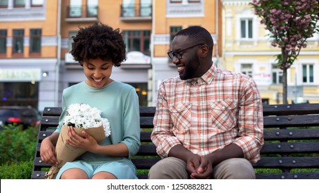 Black male giving flowers bouquet to woman sitting on bench, first awkward date
