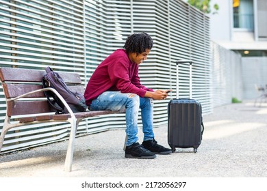 Black Male Traveler Using Smart Phone While Sitting On City Bench With Luggage