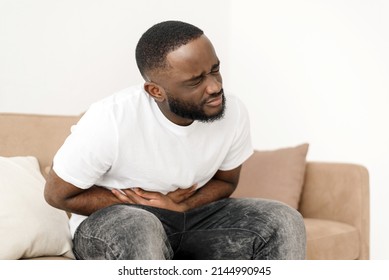 Black Man With Abdominal Pain Suffering From Painful Abdominal Cramp In Stomach Sitting On Couch At Home. Abdominal Pain, Stomach Inflammation And Appendicitis Concept