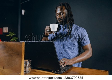 black man of African ethnicity, with dreadlocks and casual clothes, standing working with his laptop, managing his business, inside restaurant and having cup of coffee, business concept, copy space