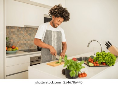 A black man in an apron joyfully slices cucumbers on a cutting board in a bright, modern kitchen filled with fresh vegetables and vibrant greens, preparing a nutritious meal. - Powered by Shutterstock