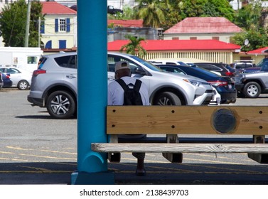 A Black Man With A Backpack Sits On A Wood Bench Facing Away While Waiting For A Bus On A Sunny Summer Day